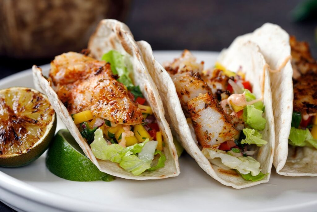 Three fish tacos filled with lettuce, mango, and diced vegetables, served with grilled lime on a white plate.