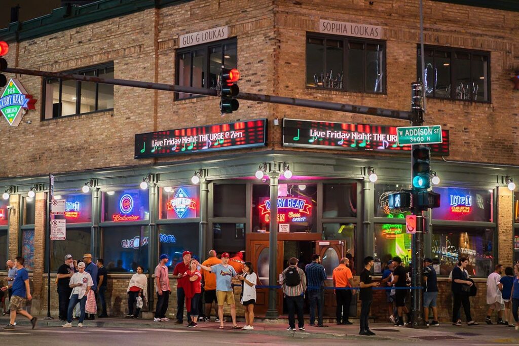 A lively street scene at night featuring a bar with neon signs and a crowd of people outside. Traffic lights and street signs are visible