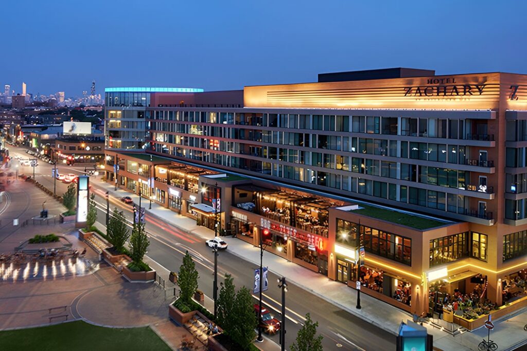 A modern hotel with illuminated signage, surrounded by city streets and greenery, captured at dusk