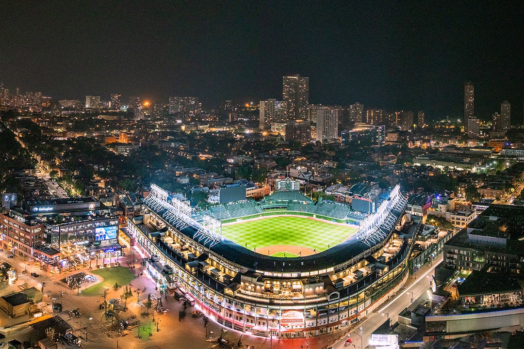 Aerial view of a brightly lit baseball stadium at night, surrounded by a cityscape with tall buildings and urban lights.
