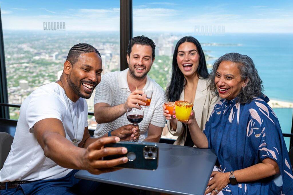 A group of four friends enjoying drinks and taking a selfie at a high-rise bar with a city view