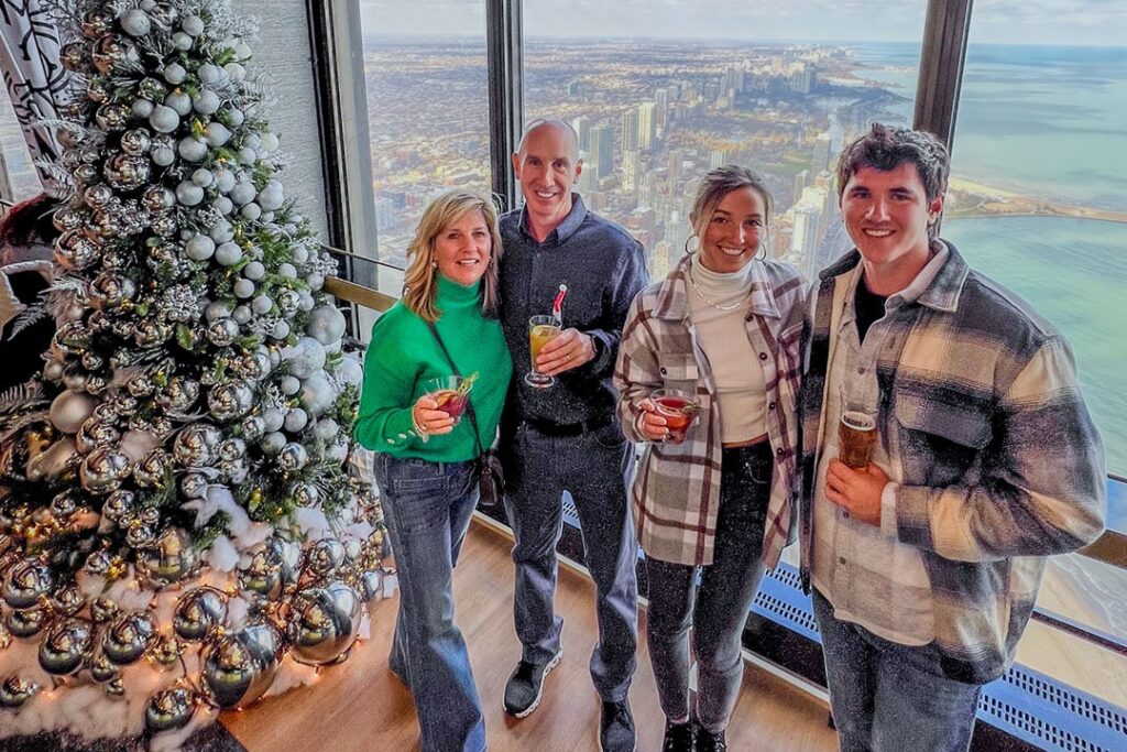 Four guests with holiday drinks beside a silver-ornament Christmas tree at 360 CHICAGO, Lake Michigan in the background.